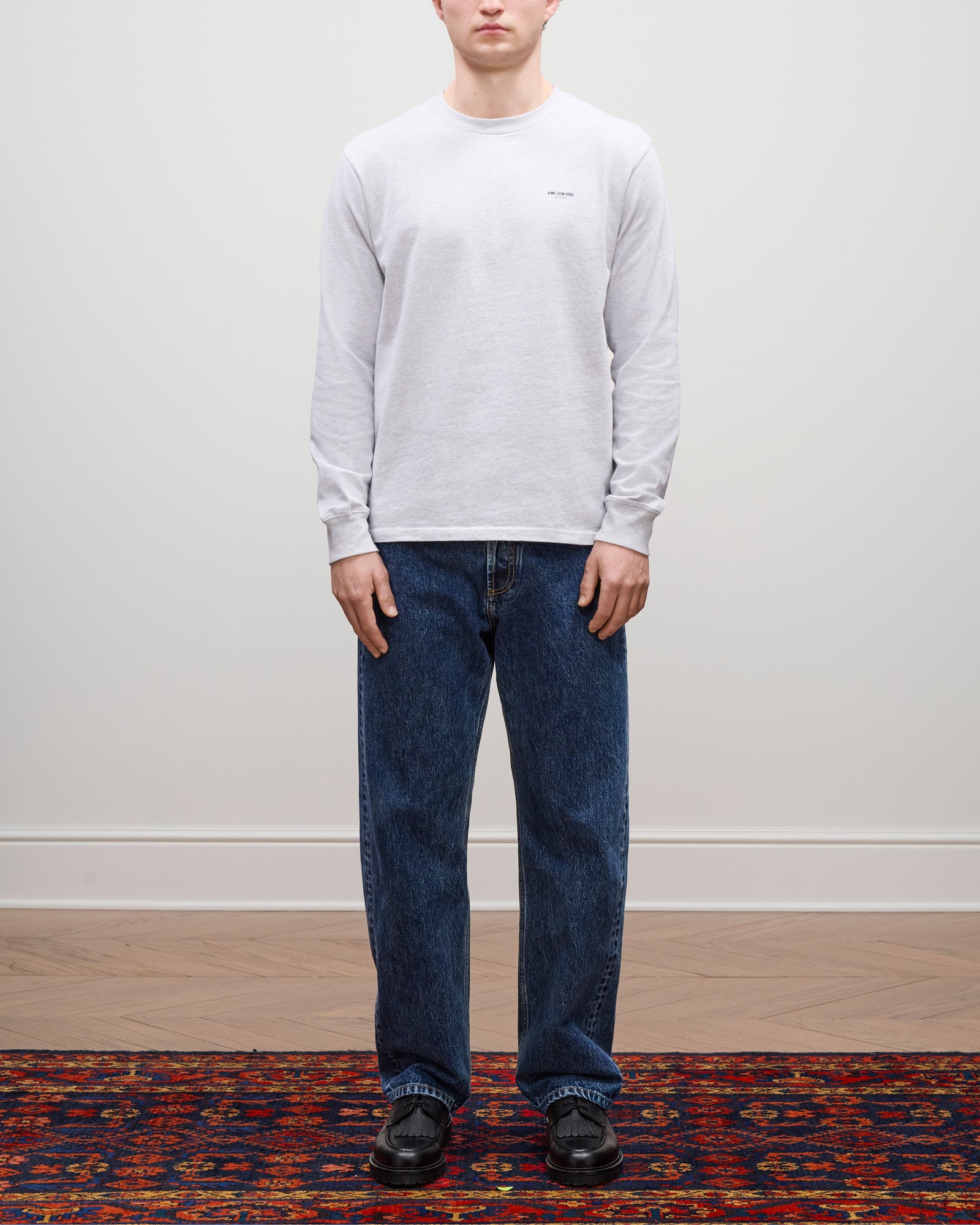 Person wears Long-Sleeve Micro Logo Tee, blue jeans, and black shoes on a patterned rug against a plain wall.