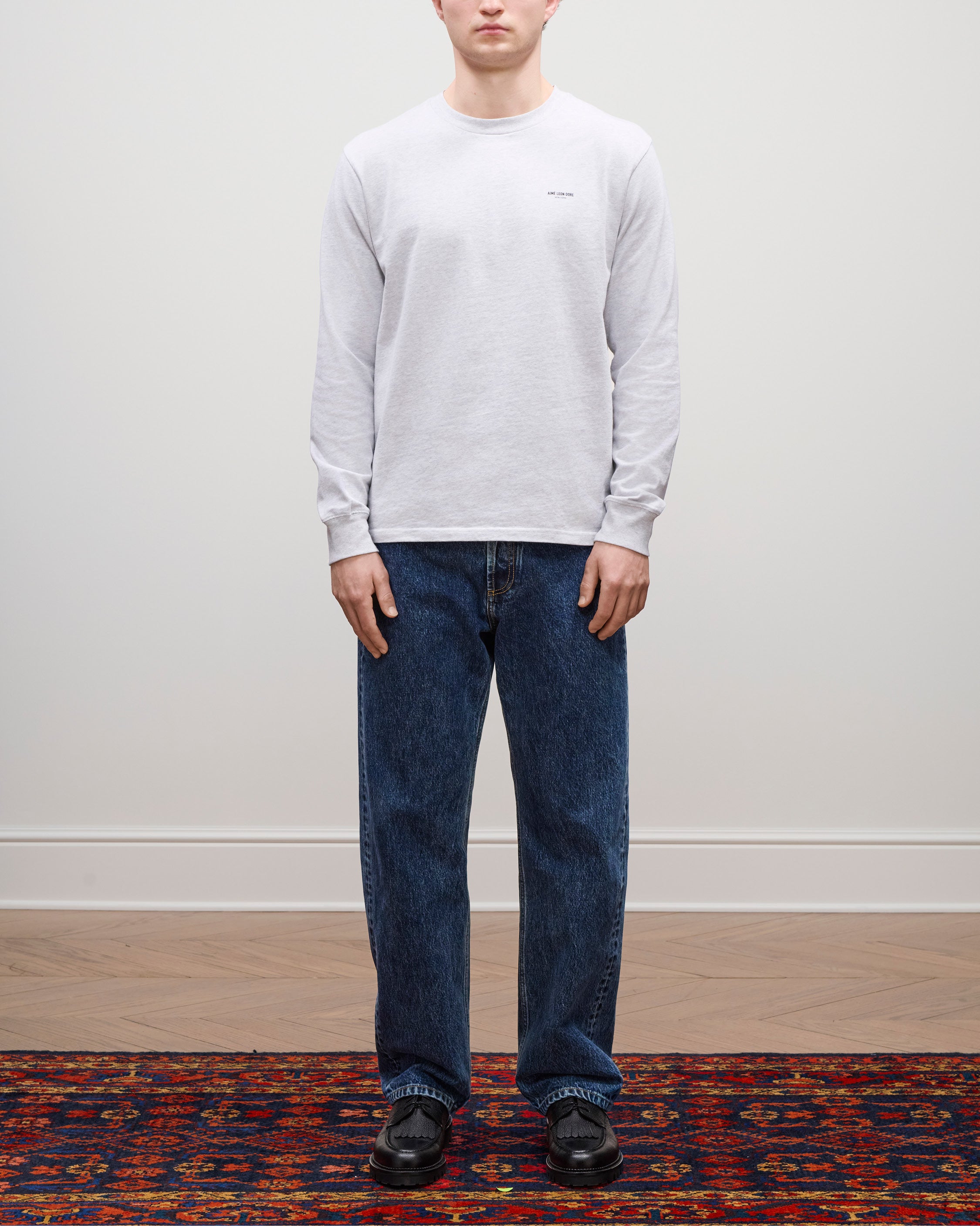 Person wears Long-Sleeve Micro Logo Tee, blue jeans, and black shoes on a patterned rug against a plain wall.