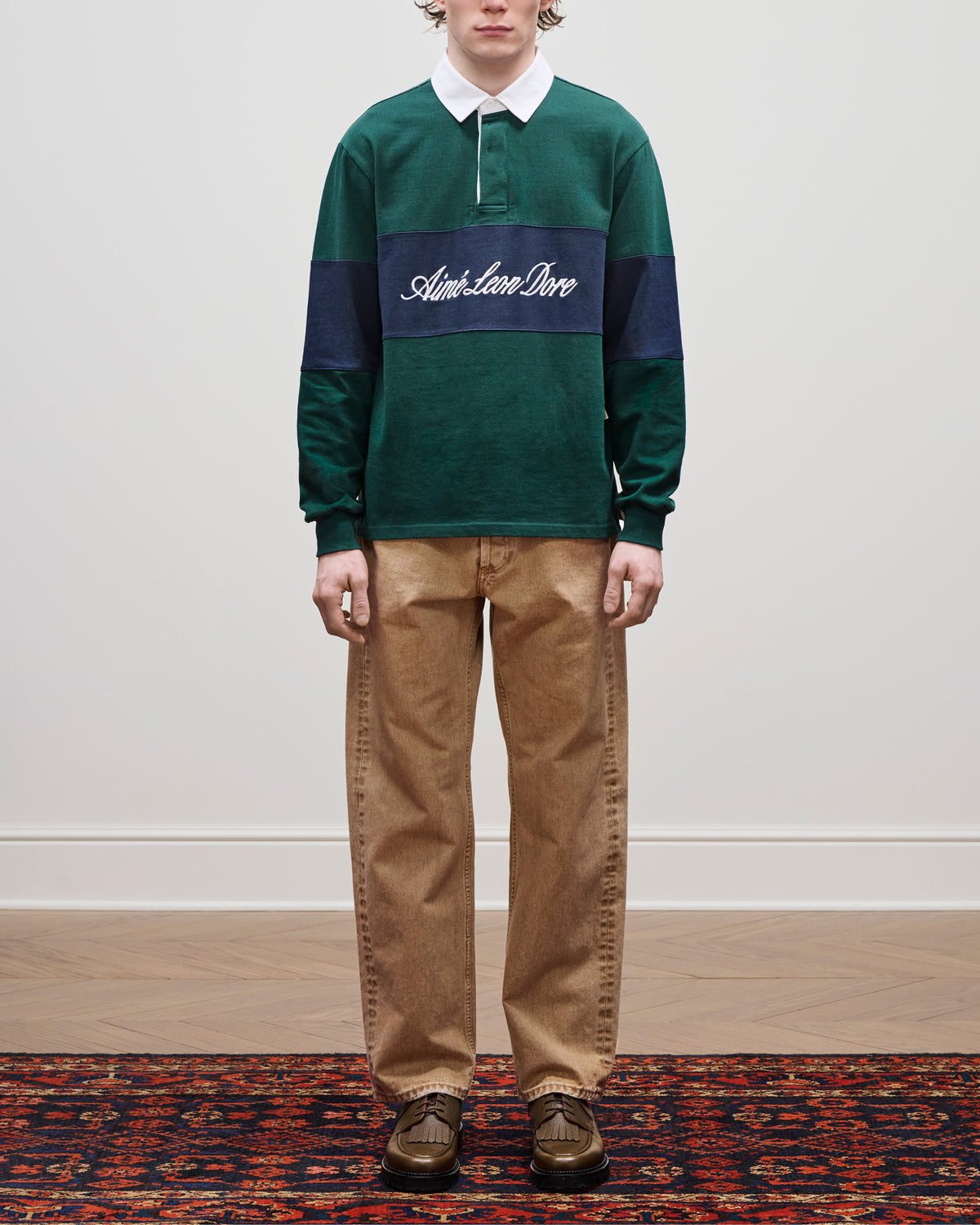 Person in green rugby shirt with Adieu Lave Dire text, Relaxed Fit 5-Pocket Denim Pant, and brown shoes on a rug.
