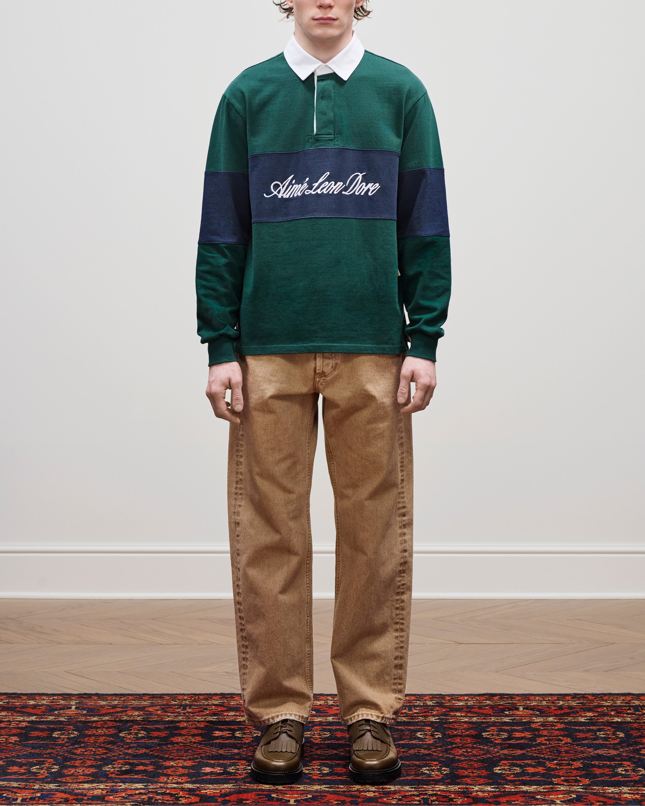 Person wears Script Paneled Rugby, tan pants, and brown shoes while standing on a patterned rug.