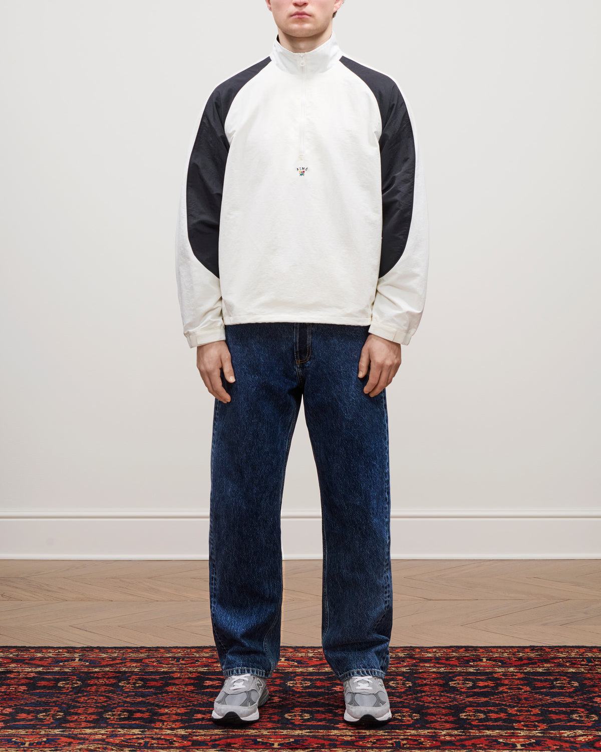 Person stands on patterned rug wearing blue Relaxed Fit 5-Pocket Denim Pant, white and black shirt, and gray sneakers.