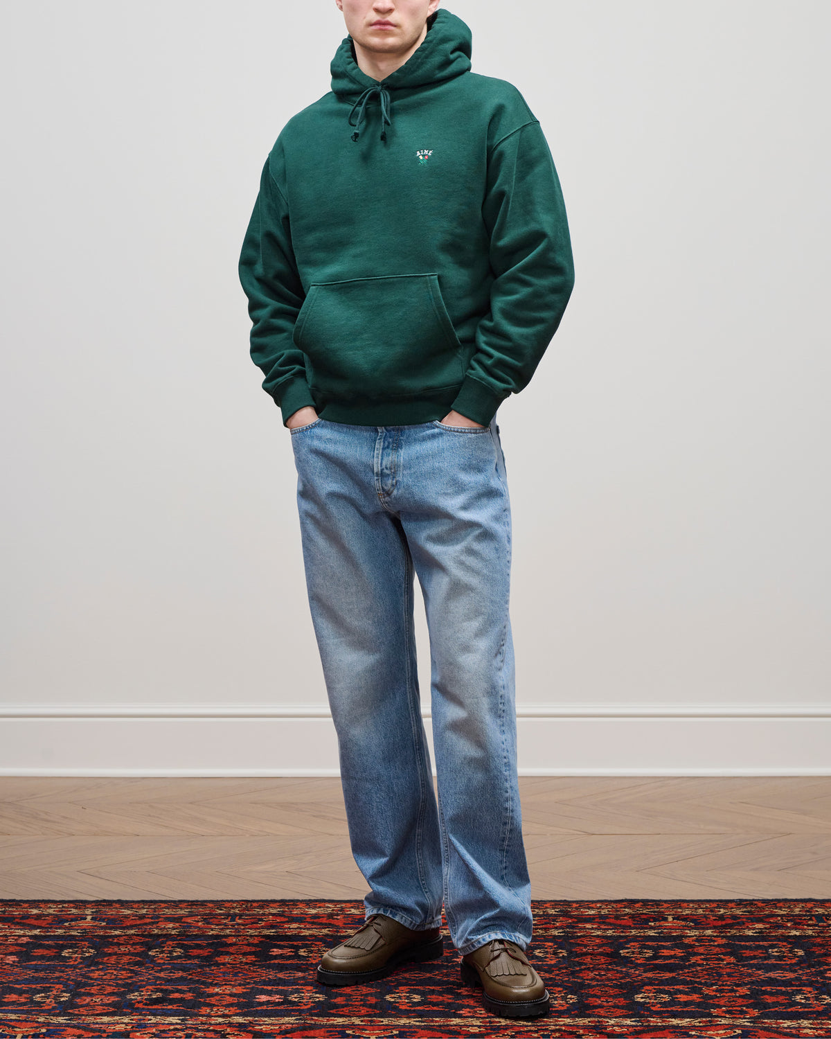 Person wearing blue Relaxed Fit 5-Pocket Denim Pant, a green hoodie, and brown shoes stands on a patterned rug.