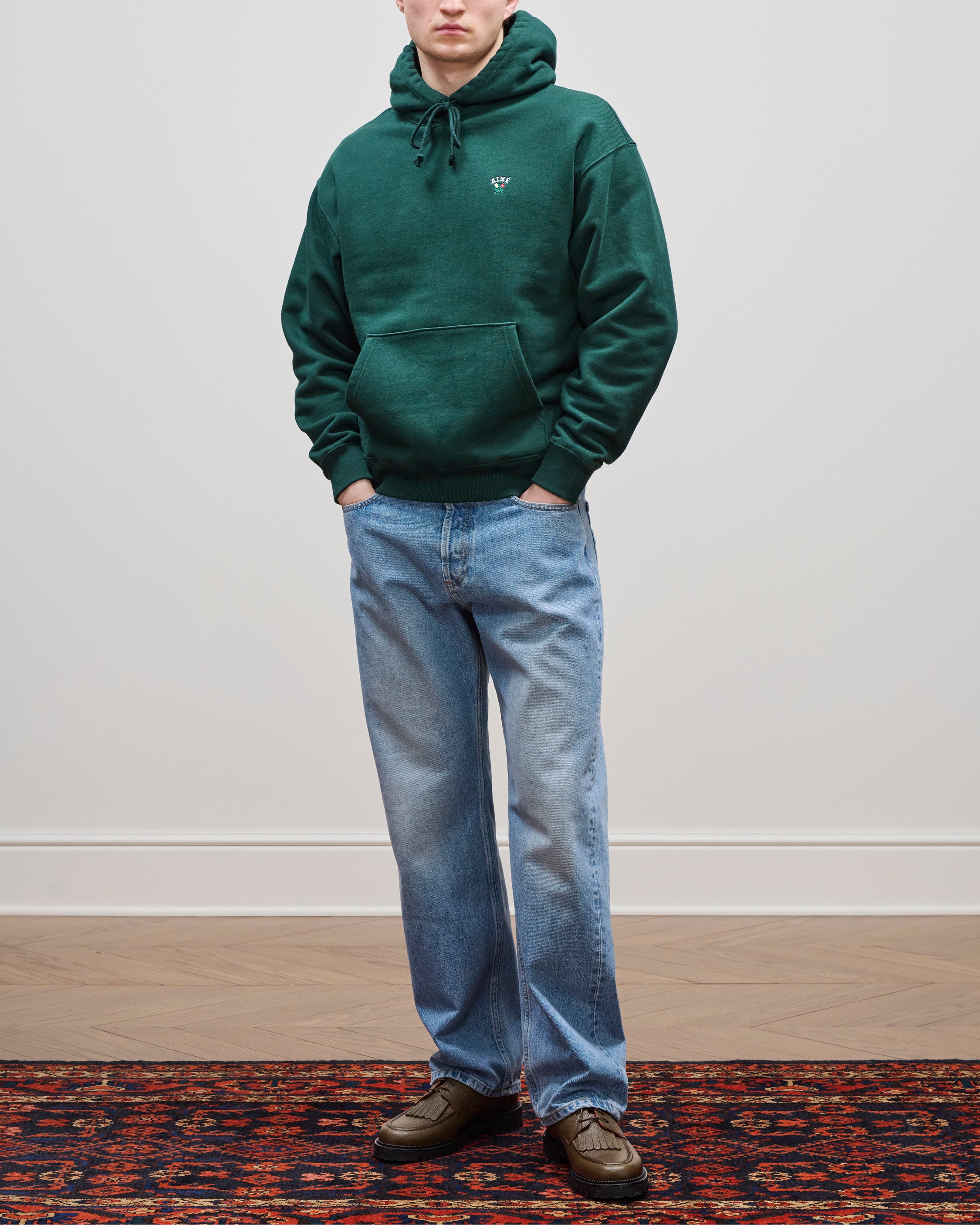 Person wearing blue Relaxed Fit 5-Pocket Denim Pant, a green hoodie, and brown shoes stands on a patterned rug.