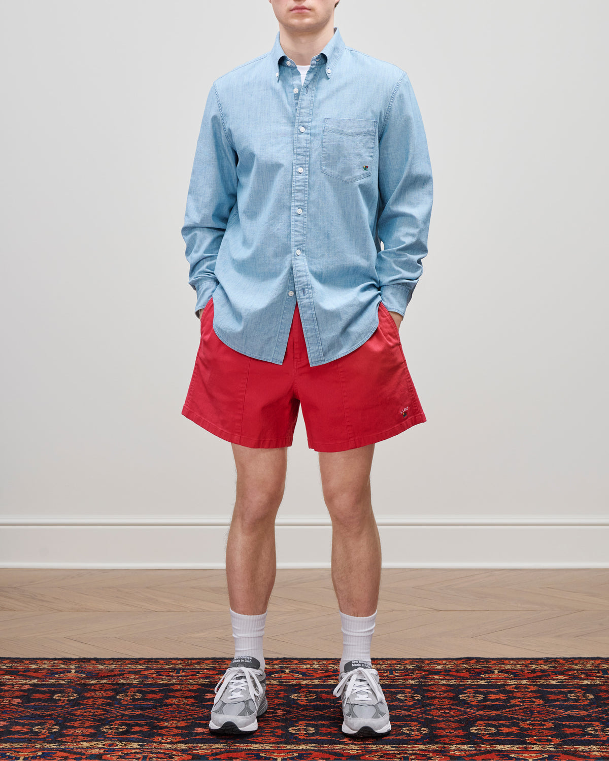 Person wearing a Crest Chambray Shirt, red shorts, white socks, and gray sneakers, standing on a patterned rug.