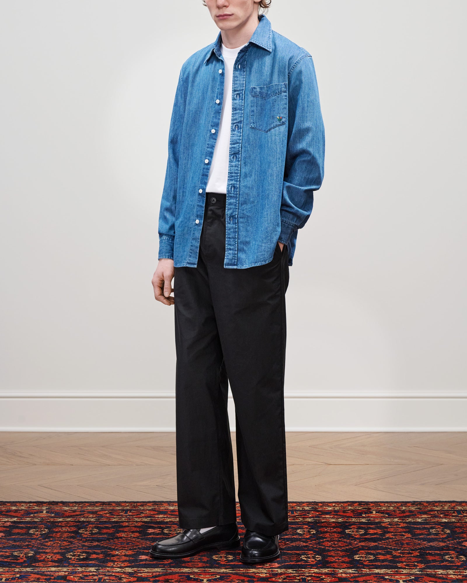 A person stands on a patterned rug wearing a Crest Chambray Shirt, white t-shirt, black pants, and black shoes.