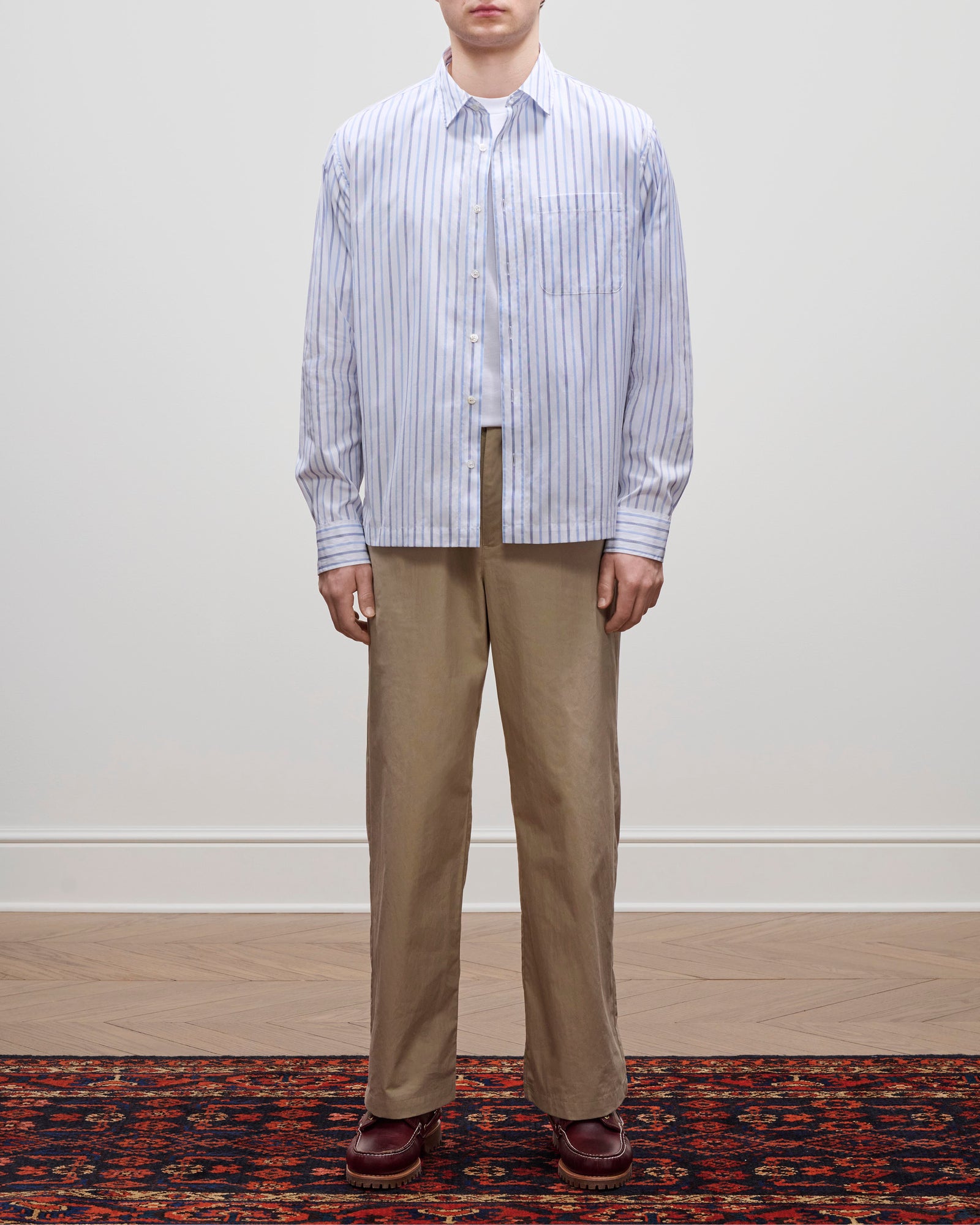 A person in relaxed cotton trousers, a light blue striped shirt, and brown shoes stands on a patterned rug.