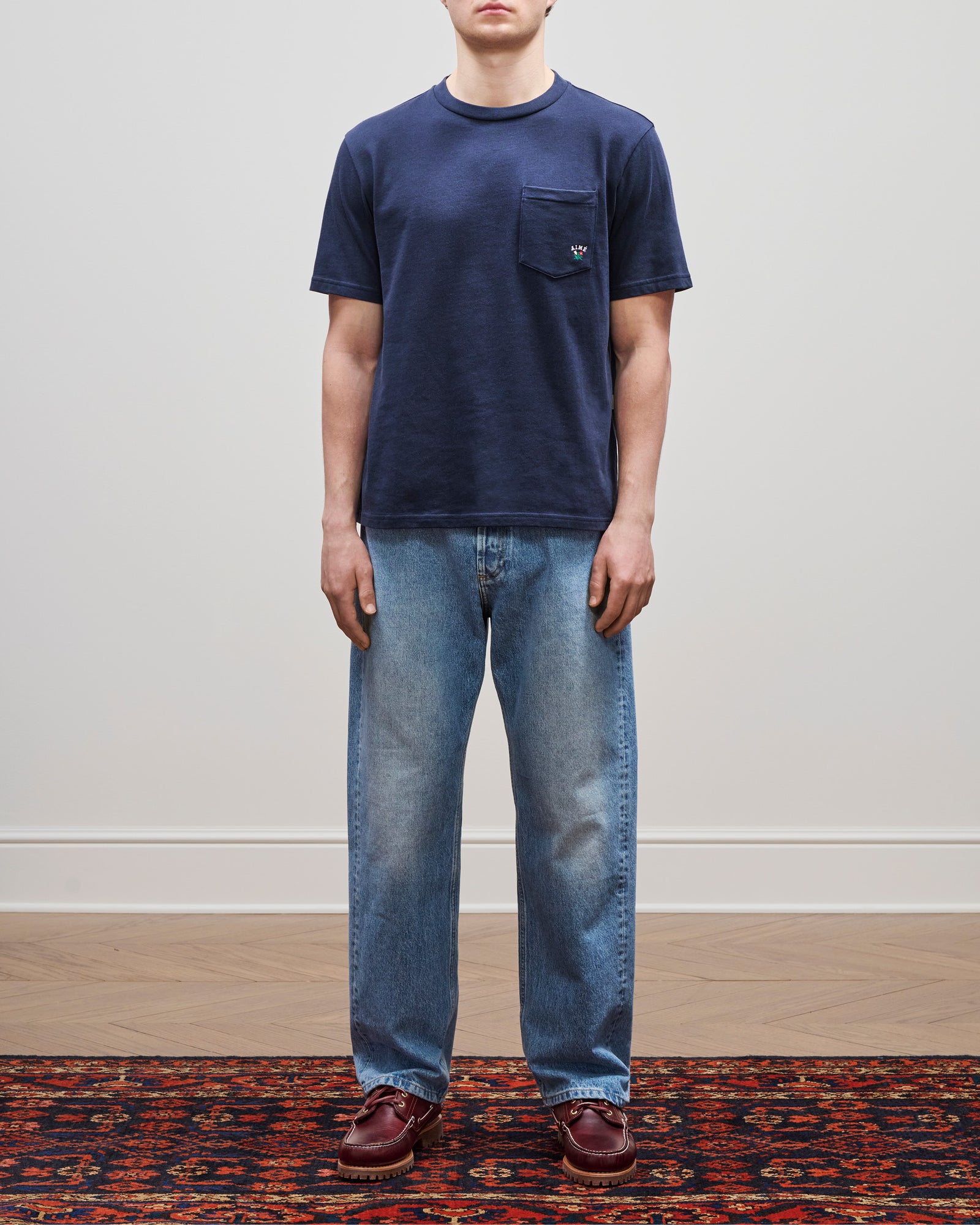 A person stands in a Crest Pocket Tee, blue jeans, and brown shoes on a patterned rug against a white wall.