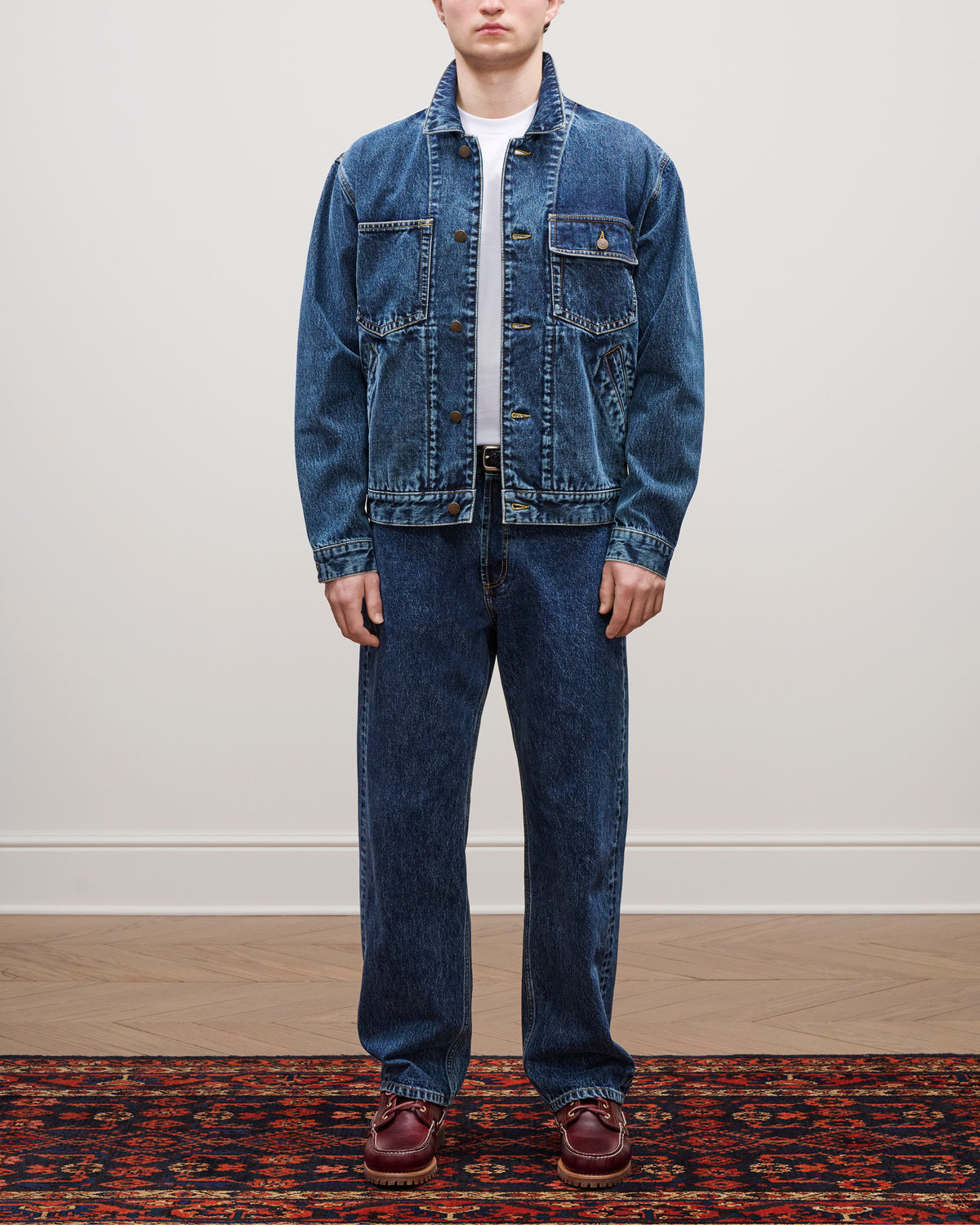 A person stands on a patterned rug wearing a Denim Trucker Jacket, blue jeans, white shirt, and brown shoes.