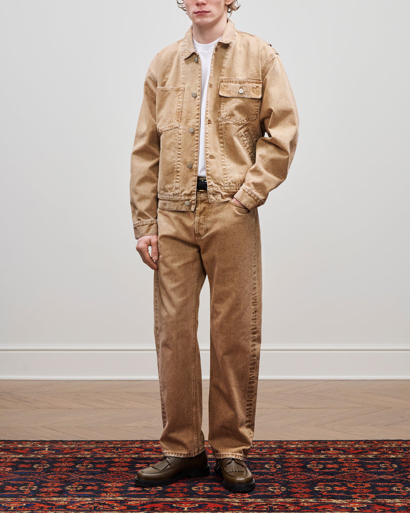 A person wears a tan Denim Trucker Jacket, pants, white shirt, and green shoes on a patterned rug by a plain wall.