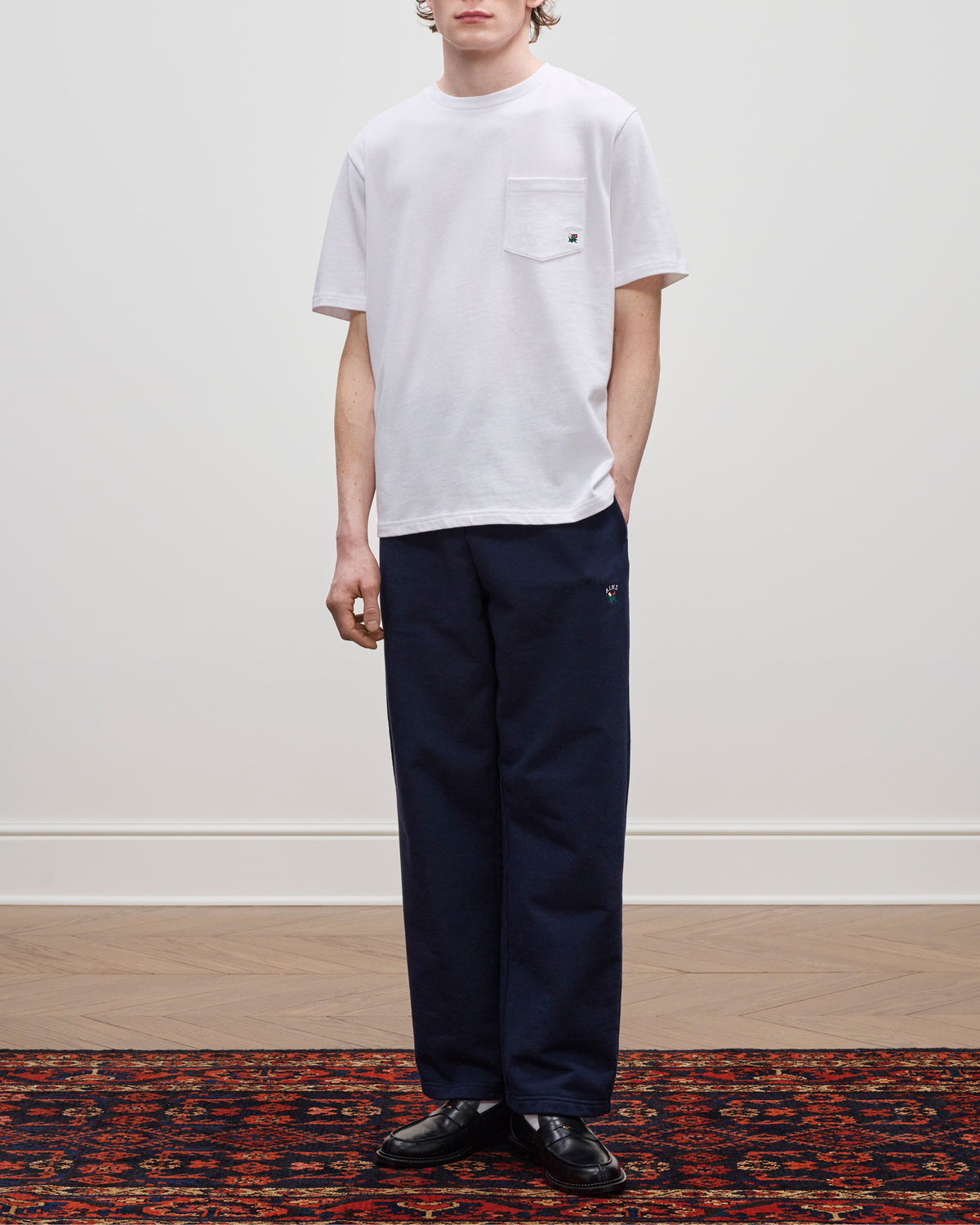 Wearing the Crest Pocket Tee in white with navy pants and black shoes, standing on a patterned rug by a plain wall.