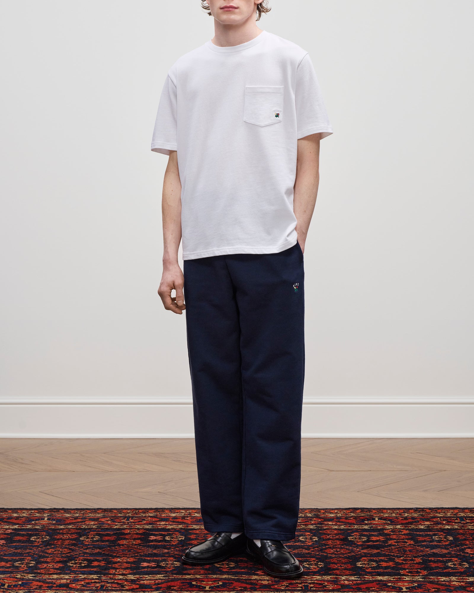 Wearing the Crest Pocket Tee in white with navy pants and black shoes, standing on a patterned rug by a plain wall.