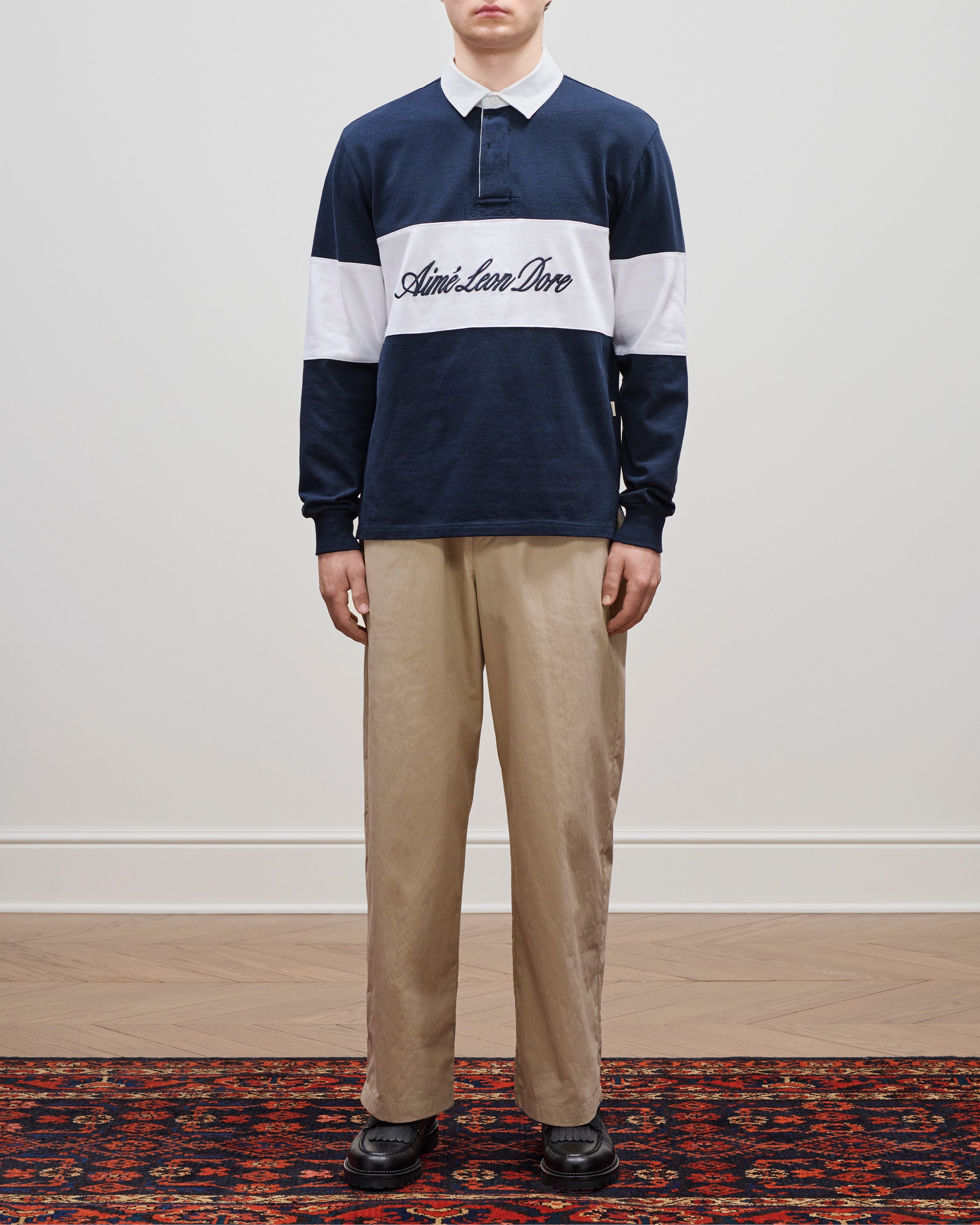 A person wears the Aimé Leon Dore Script Paneled Rugby shirt, khaki pants, and black shoes.