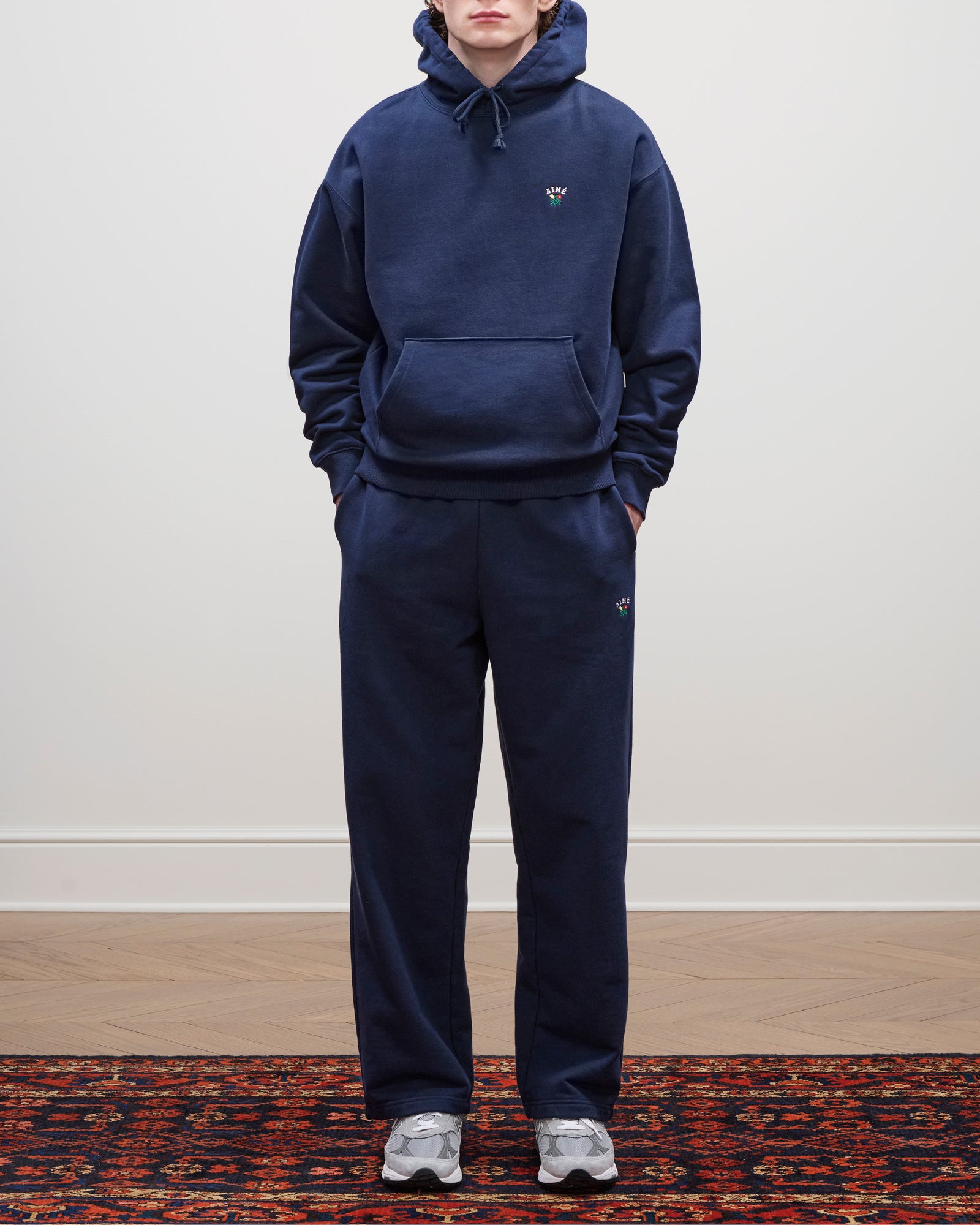 Person wears navy Crest Sweatpants, a matching hoodie, and gray sneakers, standing on a patterned rug by a plain wall.