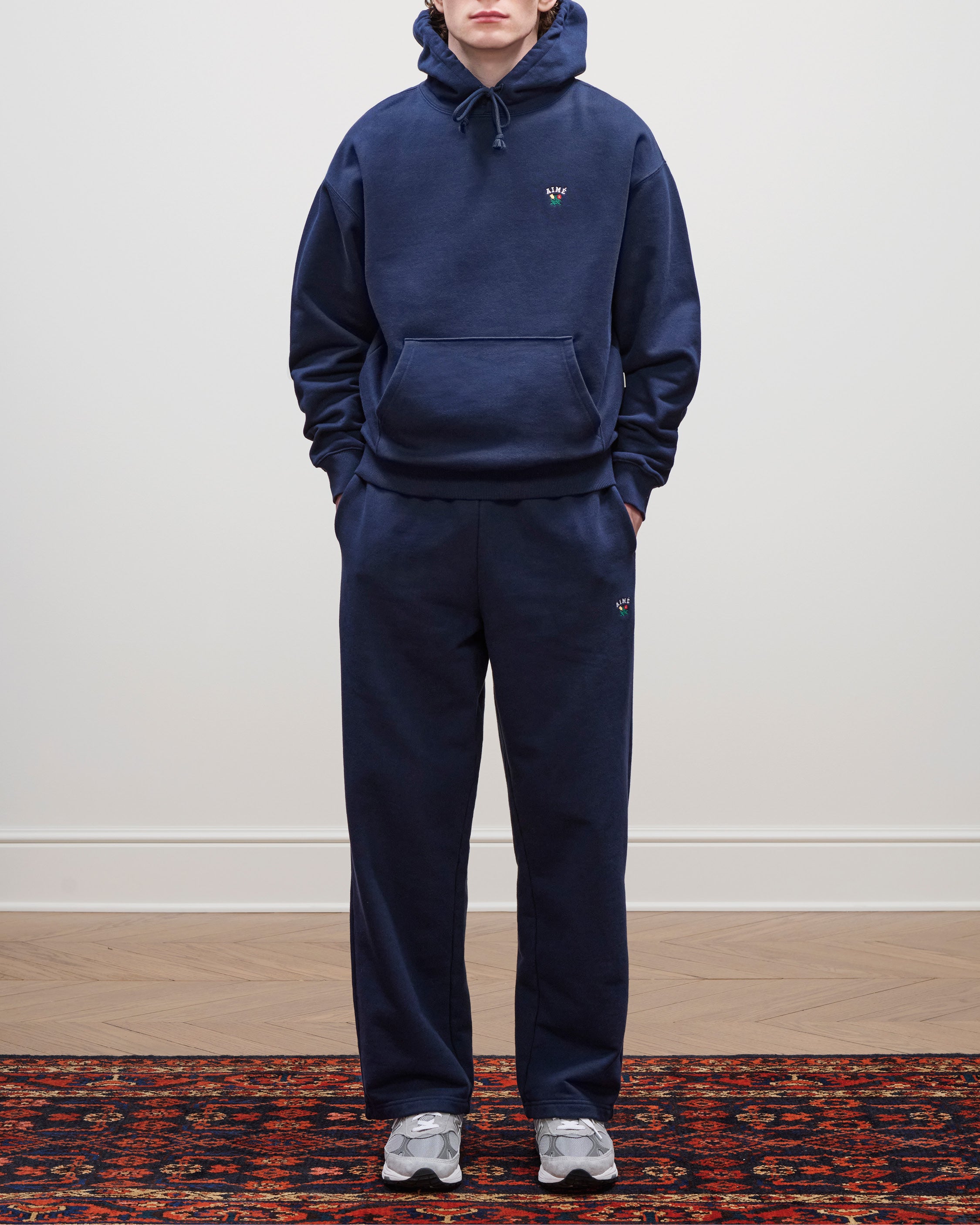 Person wears navy Crest Sweatpants, a matching hoodie, and gray sneakers, standing on a patterned rug by a plain wall.