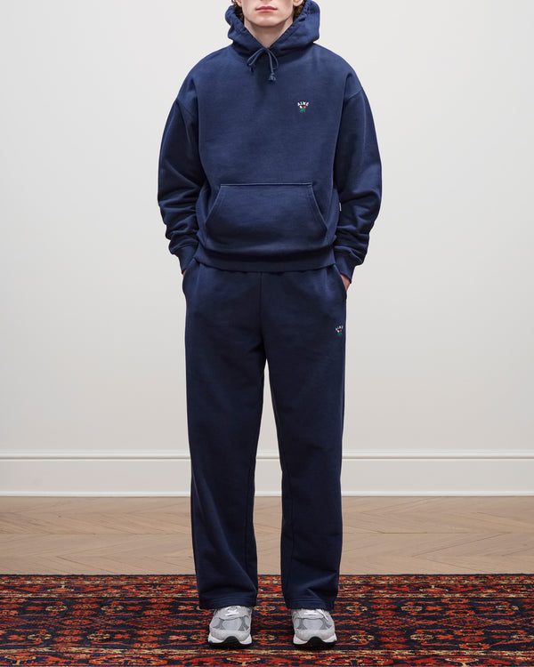 Person wears navy Crest Sweatpants, a matching hoodie, and gray sneakers, standing on a patterned rug by a plain wall.