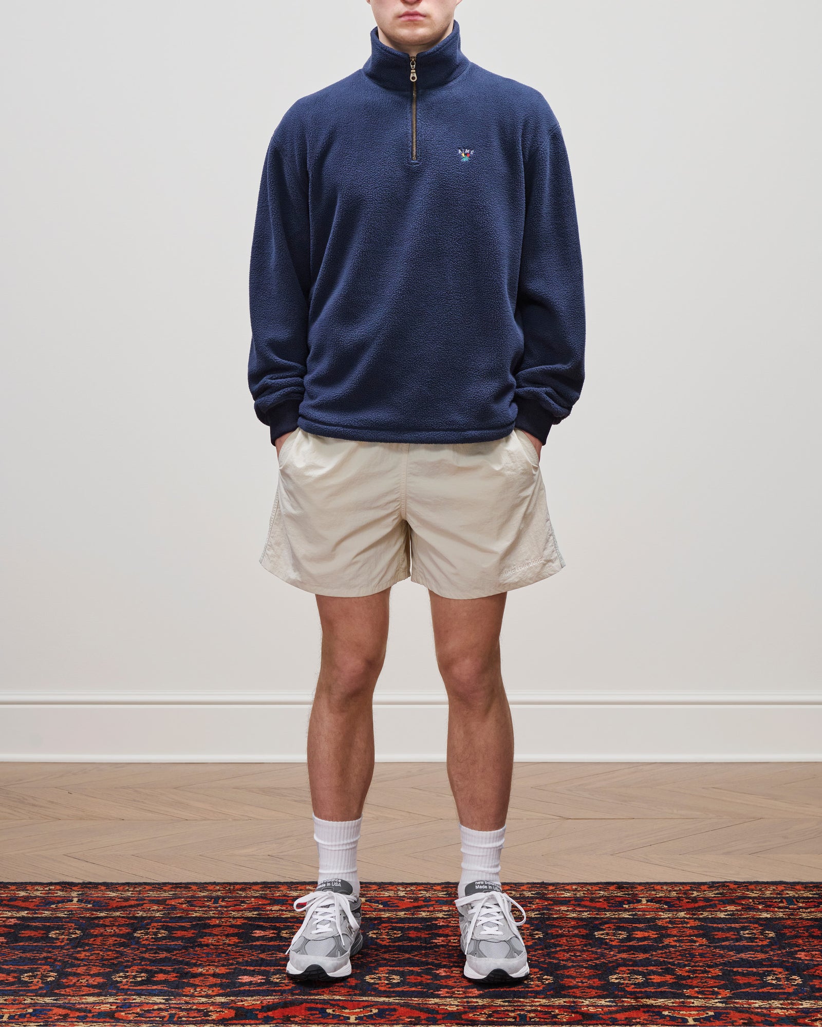 Person wearing a navy Crest Quarter-Zip Fleece Pullover, beige shorts, white socks, and gray sneakers on a patterned rug.