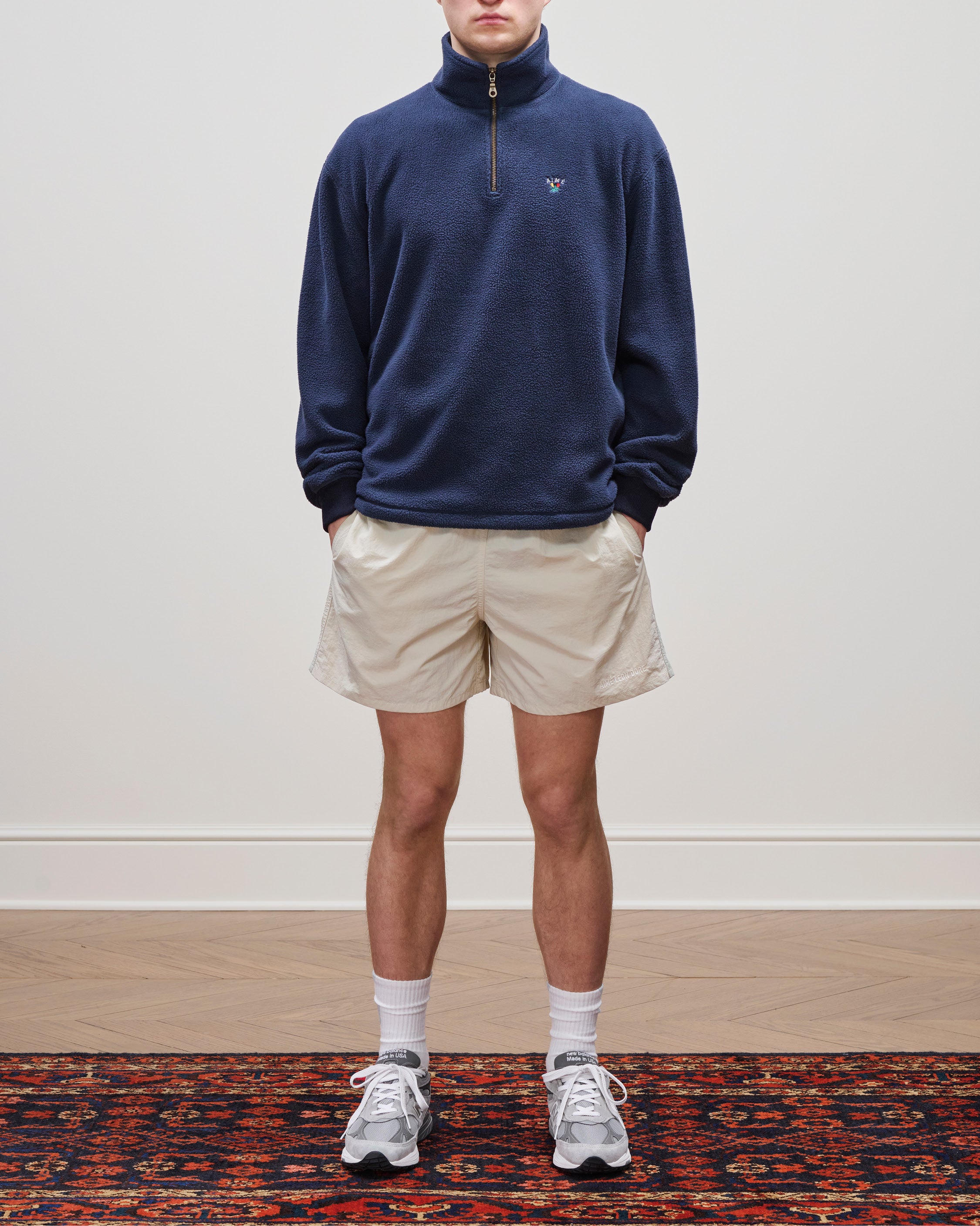 Person wearing a navy Crest Quarter-Zip Fleece Pullover, beige shorts, white socks, and gray sneakers on a patterned rug.