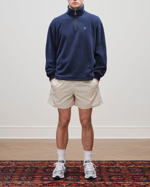 Person wearing a navy Crest Quarter-Zip Fleece Pullover, beige shorts, white socks, and gray sneakers on a patterned rug.