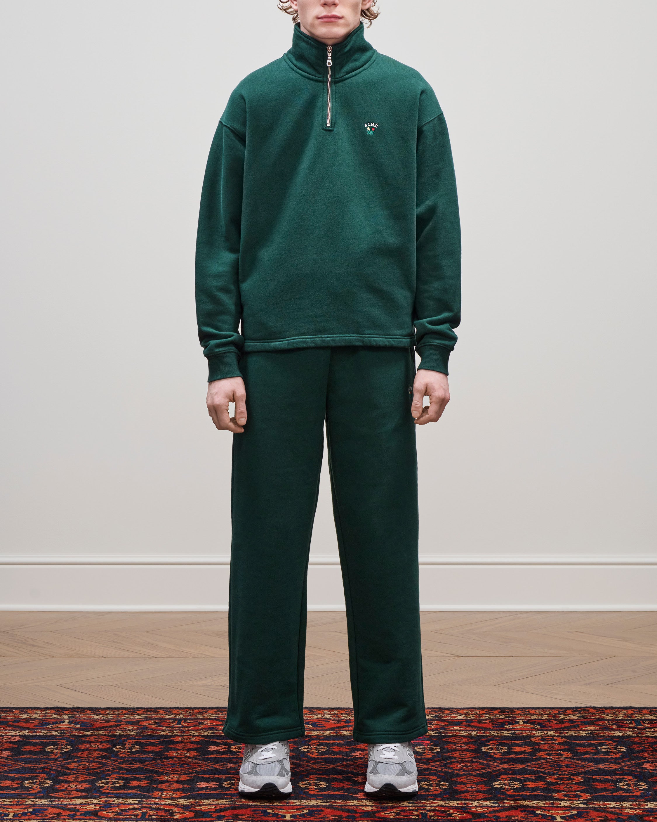 Person in Crest Sweatpants and gray sneakers stands on a patterned rug against a plain white wall.