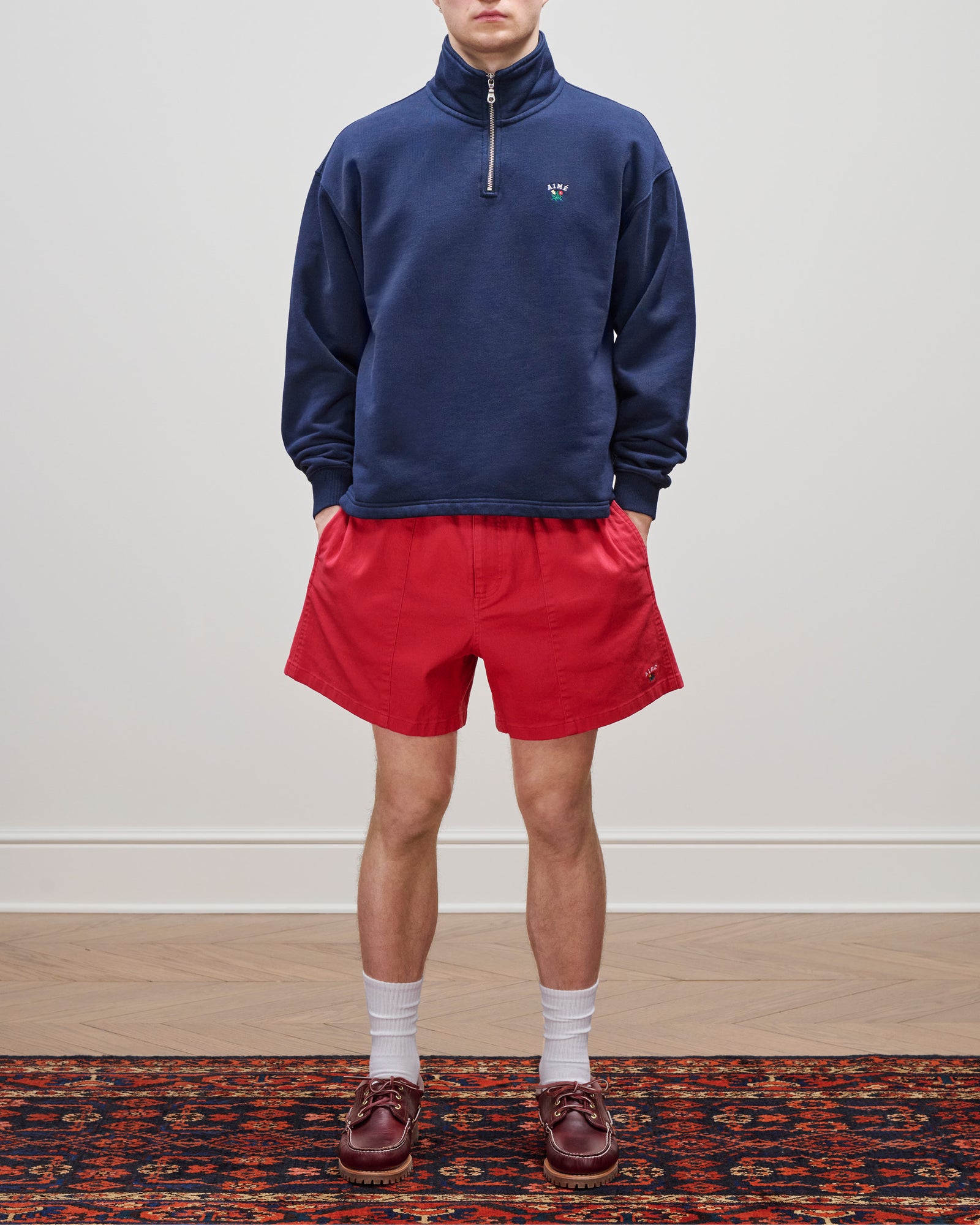 Wearing the Crest Quarter Zip Pullover in navy, red shorts, white socks, and brown loafers on a patterned rug.