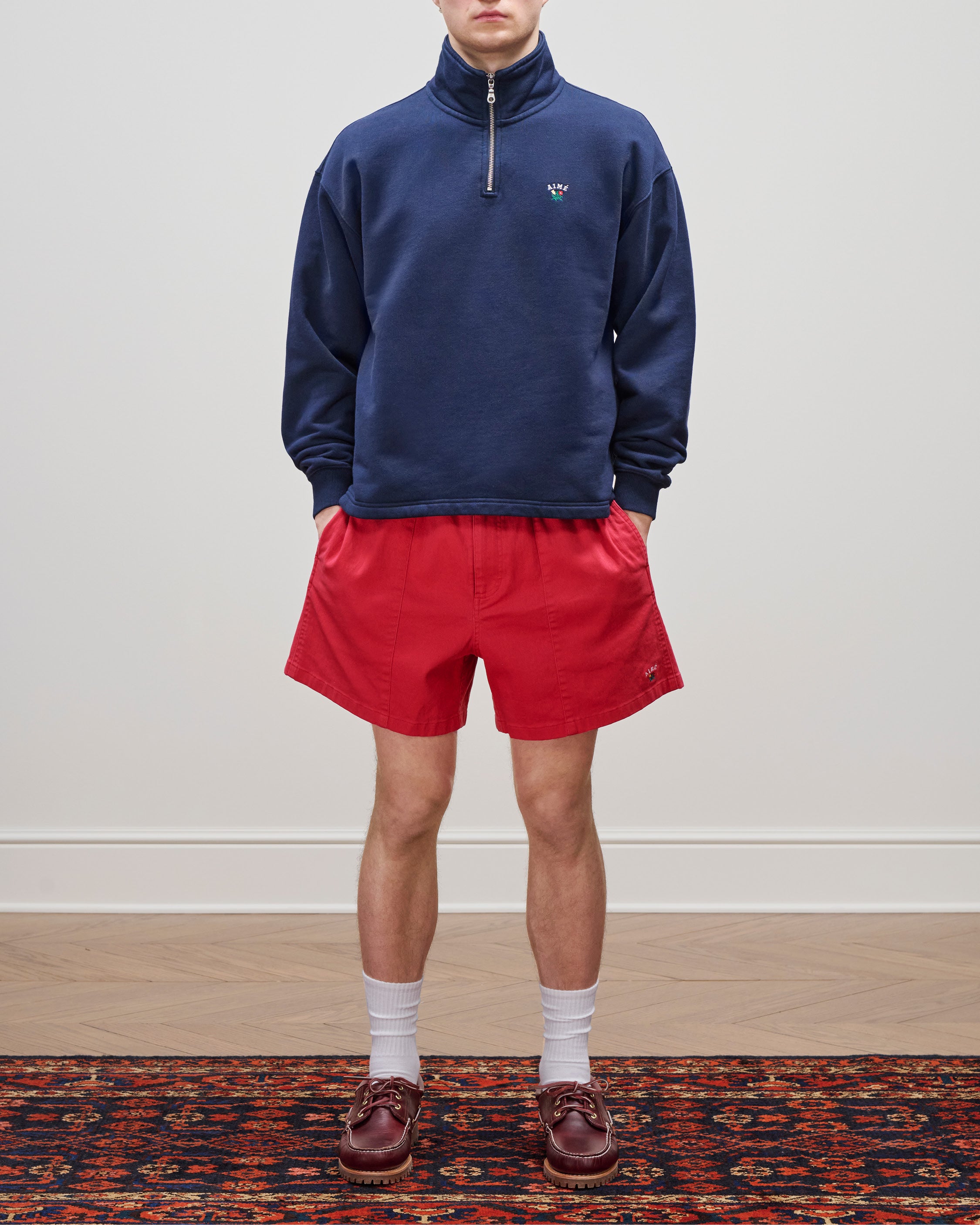 Wearing the Crest Quarter Zip Pullover in navy, red shorts, white socks, and brown loafers on a patterned rug.