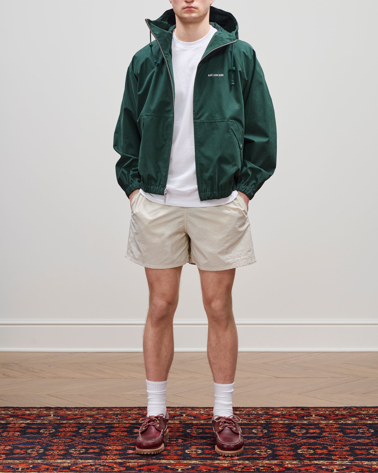 A person in a Micro Logo Lightweight Jacket, white t-shirt, beige shorts, socks, and brown shoes on a patterned rug.