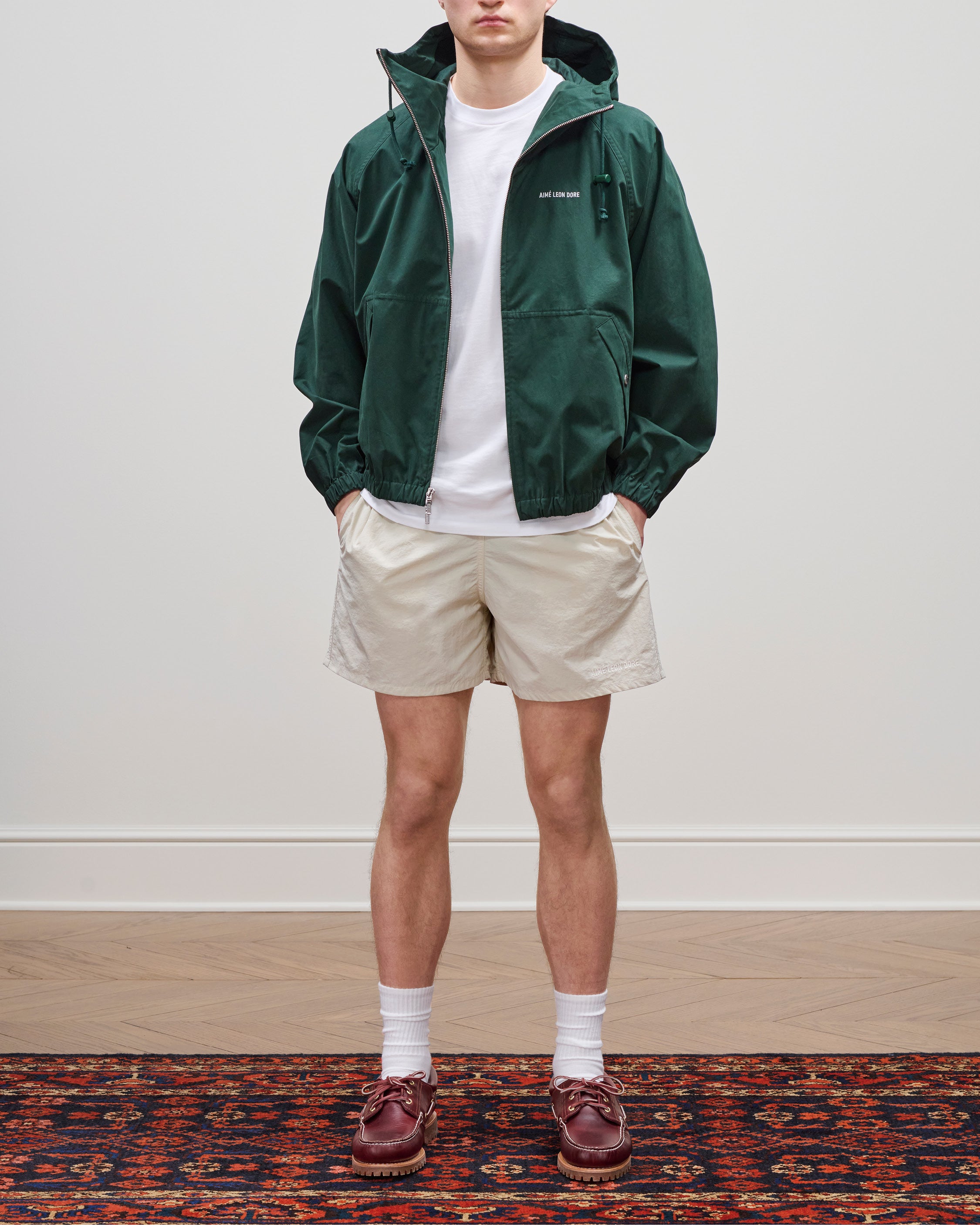 A person in a Micro Logo Lightweight Jacket, white t-shirt, beige shorts, socks, and brown shoes on a patterned rug.