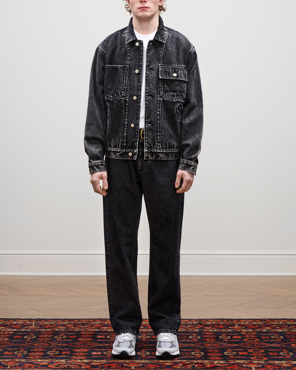 Person wearing a Denim Trucker Jacket, matching jeans, white shirt, and gray sneakers on a patterned rug.