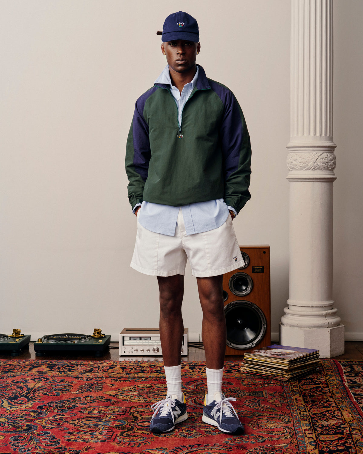 A man in a Crest Oxford Shirt, white shorts, blue cap, and sneakers stands on a patterned rug near speakers.