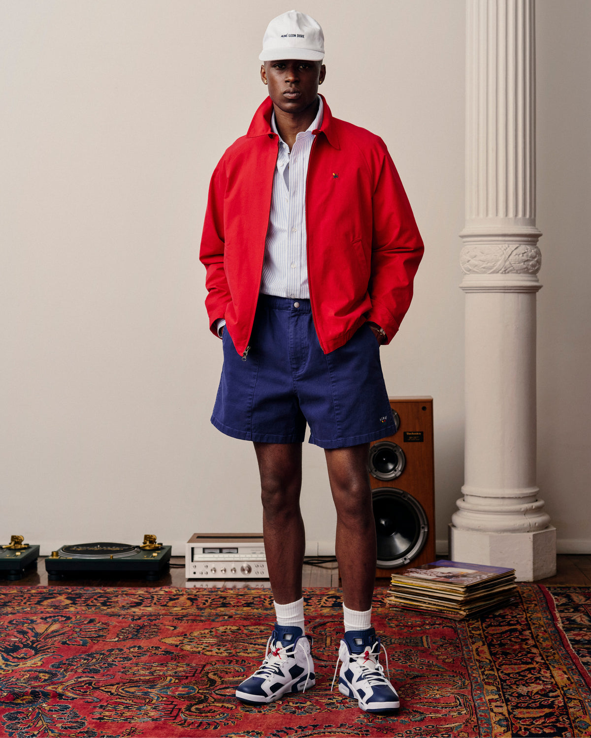 A man in a red jacket, Crest Casual Short, white hat, and sneakers stands on a colorful rug near records and speakers.