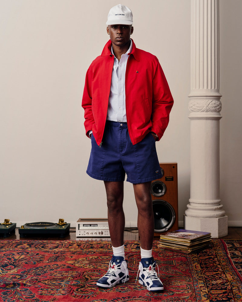 A man in a red jacket, Crest Casual Short, white hat, and sneakers stands on a colorful rug near records and speakers.