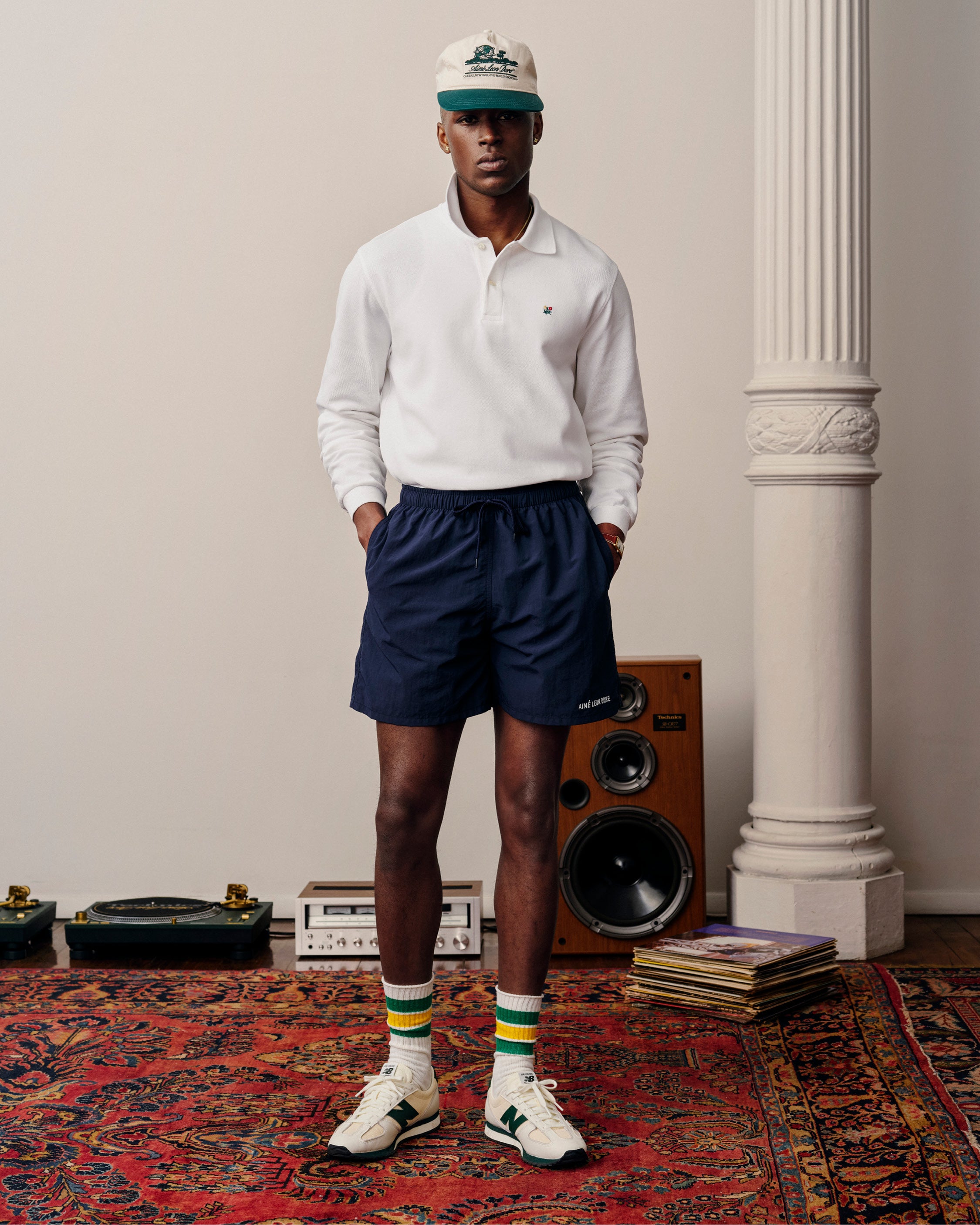 A man wears a white Long-Sleeve Crest Piqué Polo, navy shorts, cap, striped socks, and sneakers on a red rug.