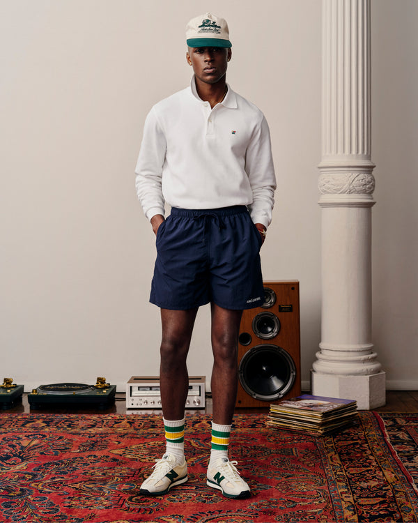 A man wears a white Long-Sleeve Crest Piqué Polo, navy shorts, cap, striped socks, and sneakers on a red rug.