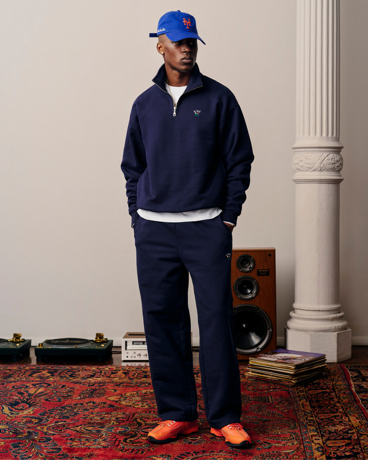 Man in Crest Sweatpants and blue Mets cap stands on a patterned rug near speakers, vinyls, and a white pillar.
