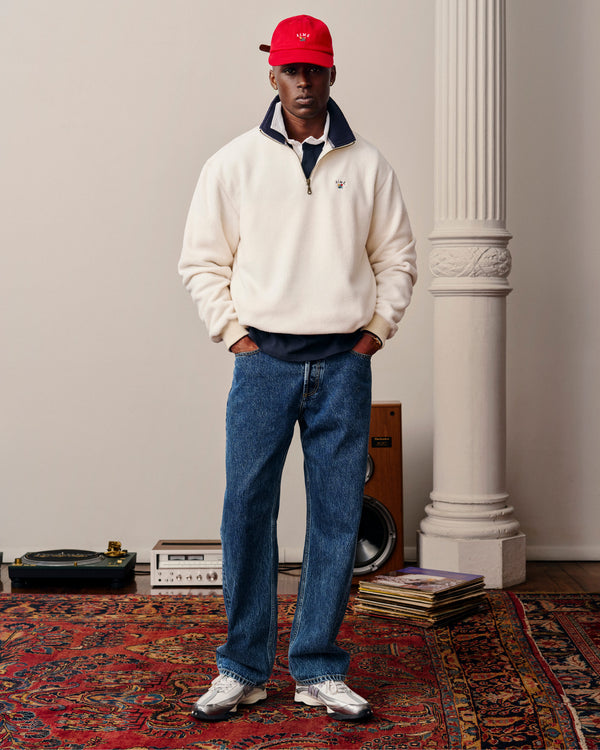 A man in a red cap, white zip sweater, Relaxed Fit 5-Pocket Denim Pant, and sneakers stands on a patterned rug indoors.