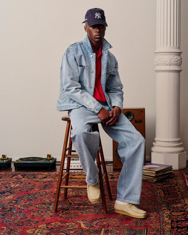 Man in Denim Trucker Jacket, jeans, red shirt, NY cap, sitting on a stool in a stylish room with a patterned rug.