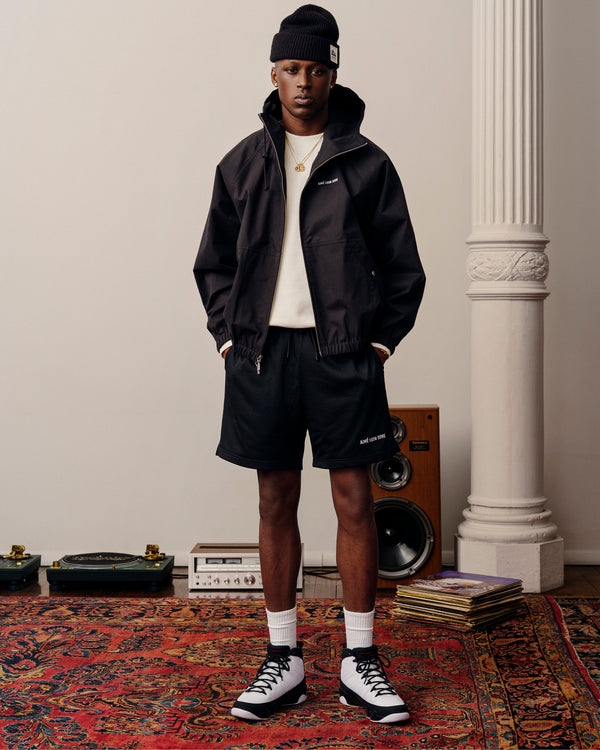 A person in a Micro Logo Lightweight Jacket, shorts, and sneakers stands on a patterned rug by speakers and records.