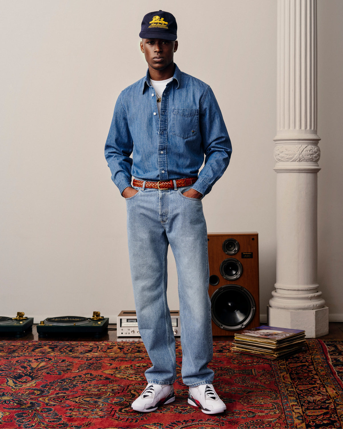 A man in ALD Straight Fit Denim stands on a red rug with speakers and records behind him.
