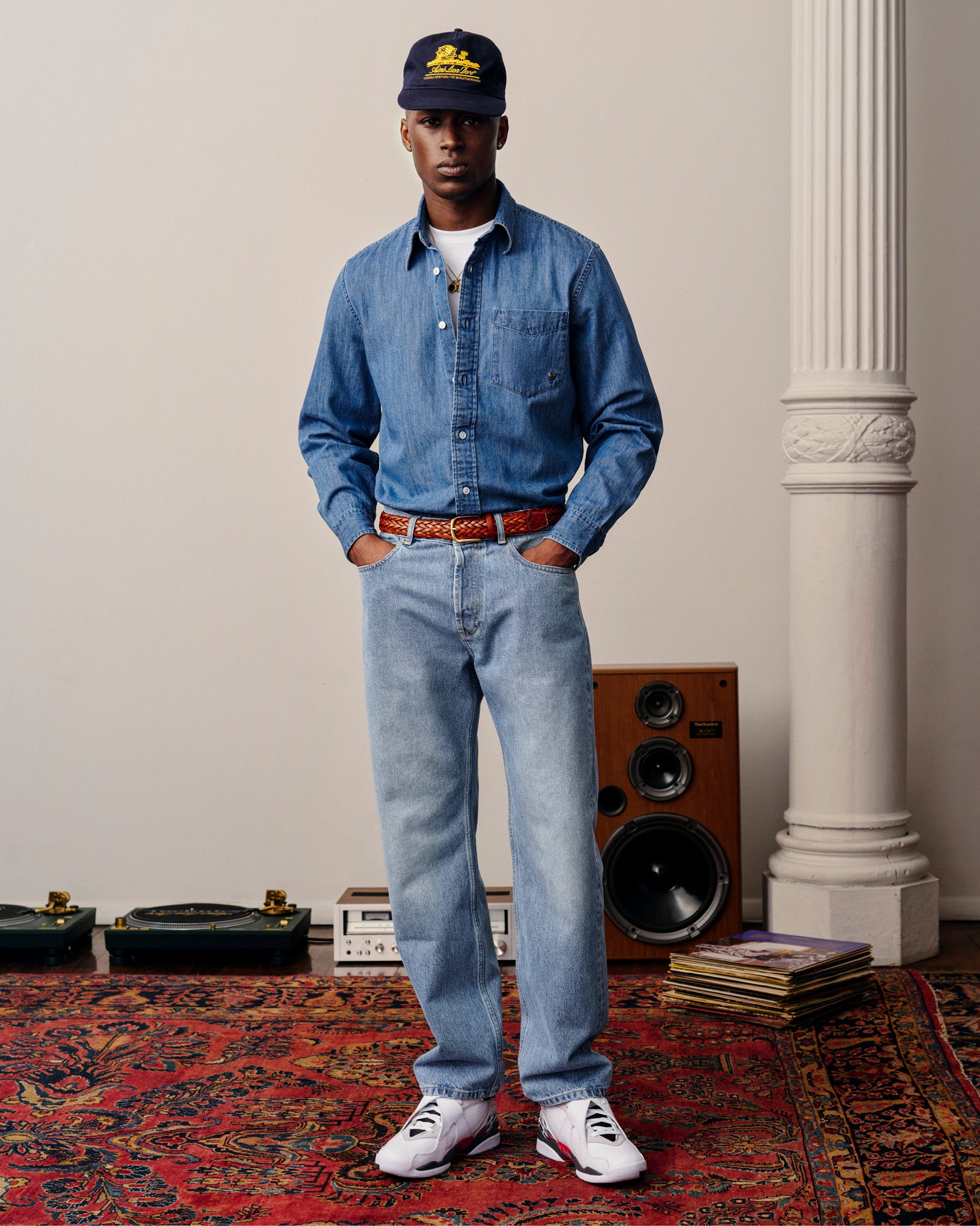 Man in Crest Chambray Shirt, light blue jeans, sneakers, and navy cap stands on red patterned rug by speakers and records.
