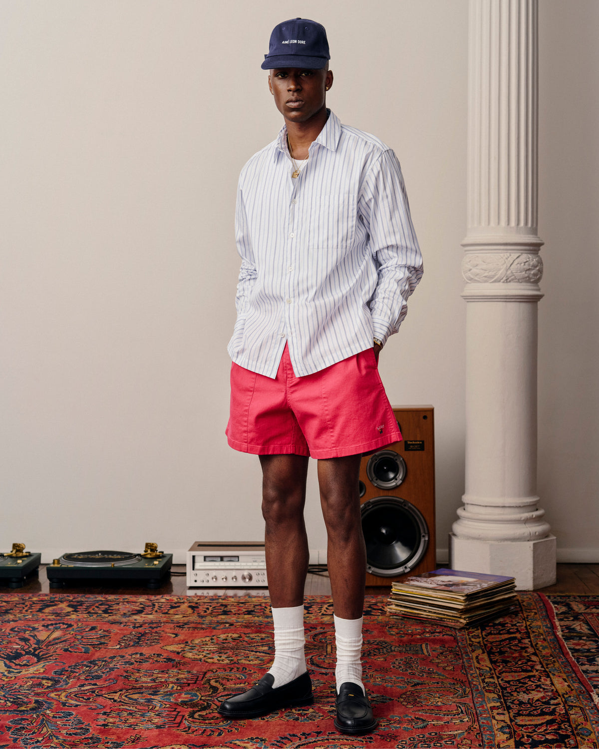 Man in Poplin Casual Shirt, red shorts, socks, loafers, and cap stands on a patterned rug near speakers and vinyl records.