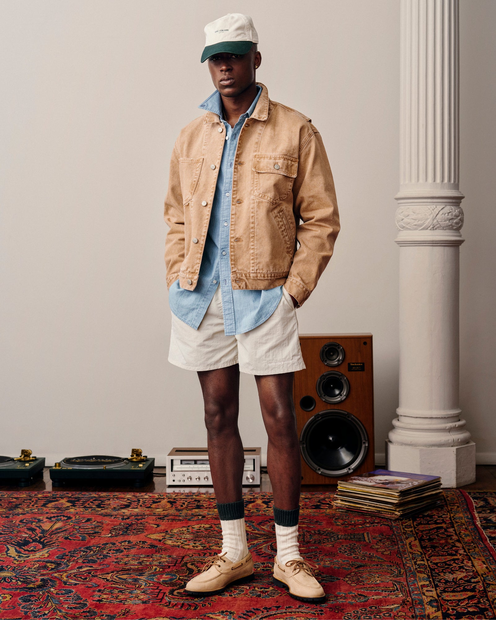 Man in a Denim Trucker Jacket, light blue shirt, beige shorts, cap, and boots stands on a rug by speakers and records.
